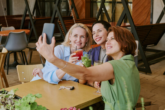 Three middle aged Caucasian women sitting at table smiling and holding cocktails while taking selfie with smartphone, showing friendship and leisure time in modern cafe setting - Powered by Adobe