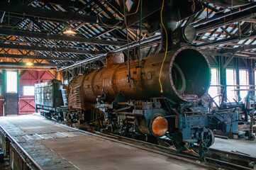 A vintage steam locomotive is showcased inside a spacious train shed, illuminated by natural light. The weathered engine rests on the tracks, surrounded by the rustic interior and wooden beams.