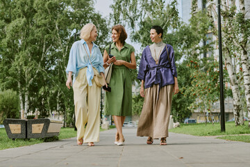 Three middle aged women walking together outdoors in park, smiling and talking, trees and city buildings visible in background, casual summer clothing