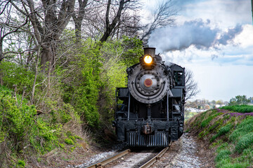 A vintage steam locomotive powers along train tracks surrounded by vibrant greenery and blooming flowers.