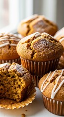 A delicious pile of freshly baked pumpkin muffins, some with powdered sugar and others drizzled with white frosting, arranged near a window letting in natural light.