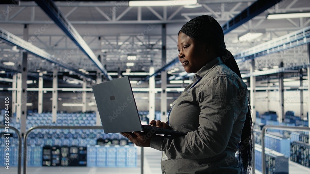 Poster it expert monitors server farm activity using laptop while walking in facility. african american wom