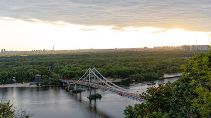 Pedestrian Bridge over the Dnipro River in Kyiv at sunset, surrounded by lush green forest, sandy beaches, and distant modern city skyline under a dramatic evening sky. 18.08.2025