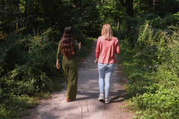 Two women walking along a forest path, photographed from behind. One wears a green outfit, the...