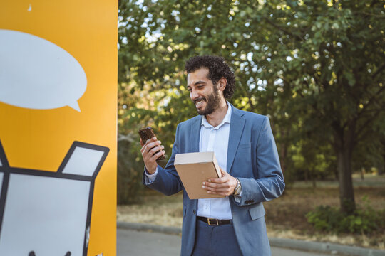 Entrepreneur using smart phone and holding package next to automated parcel locker