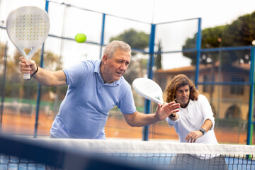Focused aged man playing friendly paddleball match on outdoor summer court. Senior people sports...