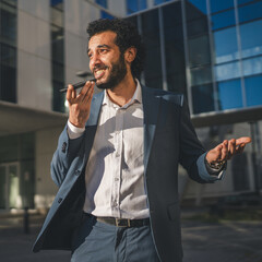 Smiling businessman sending a vocal message on his phone in front of modern office buildings