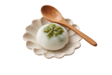 A round, light-colored confection, possibly mochi, rests on a scalloped, cream-colored dish.  A wooden spoon is positioned next to it.  Delicate, green leaf-like markings are on the top