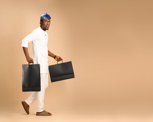 An African man in white traditional attire and blue cap walks in a studio holding two large black...