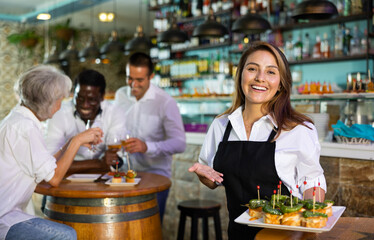 Cheerful smiling female waiter holding served tray meeting visitors at comfortable bar