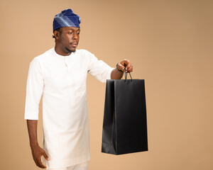 A Nigerian man dressed in traditional white attire holds a large black shopping bag in a studio...
