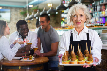 Portrait of senior female waiter inviting clients to taste plate of pinchos in cosy pub