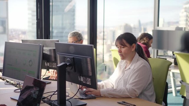 Zoom in yo a woman working at computer stations in a bright modern office with city views, combining focus and collaboration while analyzing data and discussing tasks in a shared workspace.