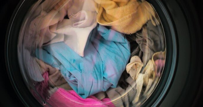 Colorful textiles spinning and tumbling inside front load washer drum demonstrating thorough cleaning process with water and detergent during typical domestic chore. Washing clothes in washing machine