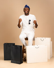 A Nigerian man in white native attire and blue fila sits on a stool, smiling and raising fists in...