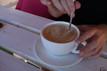 A close-up shot shows a hand stirring a creamy latte in a white cup on a rustic wooden table,...