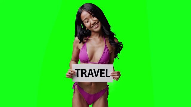 Real video of a smiling brunette woman in a pink bikini holding a white sign with the word &ldquo;TRAVEL&rdquo; in bold black letters. The model is isolated on a bright green chroma key background, perfect for ea