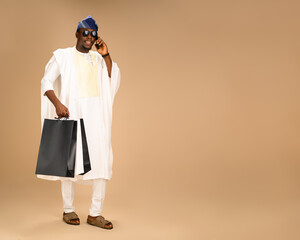 Fototapeta premium Nigerian man in agbada with blue cap and sunglasses holding shopping bags and talking on mobile phone, festive consumer lifestyle concept for retail, ecommerce, and modern holiday shopping.