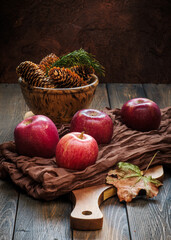 Rustic wood plank table with Fall red apples on brown gauze fabric and cutting board, with bowl of pinecones. Autumn seasonal scene