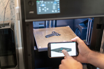 User adjusts a 3D printer's settings using a smartphone to ensure precise printing of a detailed design in a home workspace filled with tools and materials.