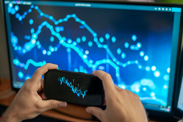 A person shows a smartphone screen with a stock graph while a larger digital display illustrates market trends behind them. The setting highlights financial analysis.