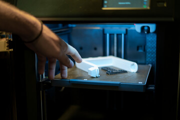 A 3D printer is in action, producing a white item with intricate designs on a build plate in a well-lit workshop, showcasing advanced printing technology.