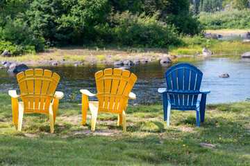 Fototapeta premium Three plastic Adirondack chairs, two yellow and one blue, on the river banks. There are lush green trees and shrubs along the rocky, flowing river's edge. The garden chairs sit on lush green grass.