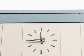 An exterior clock on a light cream coloured metal background with long grey arms. The time on the...
