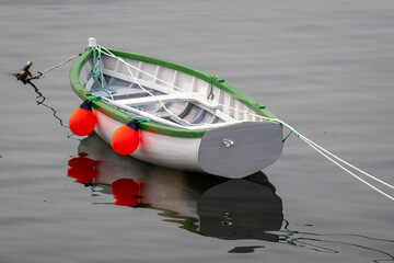 A white wooden traditional dory or small fishing vessel with green trim floating on a smooth water surface. The dory has wooden oars and a rope. The smooth water is reflecting the image of the boat. 