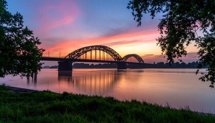 A serene riverside view at dawn, showcasing a magnificent arched bridge reflecting in the calm water under a vibrant sunrise sky.