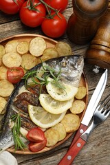 Tasty roasted dorado fish served with potatoes, tomatoes and lemon on wooden table, flat lay. Homemade seafood dish