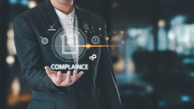 A businessman in a suit presents a glowing digital interface with the word compliance and icons representing rules and regulations. The background is a modern office.