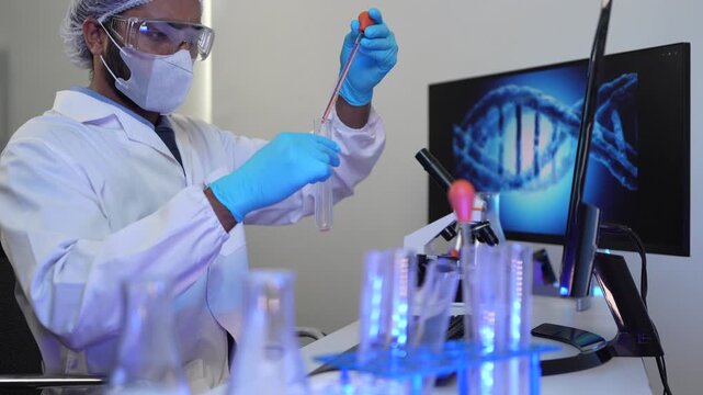 A medical researcher in a genetics lab uses a microscope to examine a sample. The DNA image on his computer screen relates to his work on disease research and diagnostics.