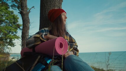 Young woman sits peacefully against tree in coastal area, holding rolled-up yoga mat and backpack. Serene mood embodies contemplation amidst nature.