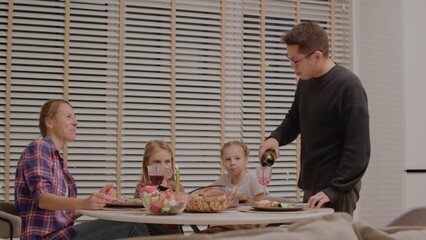 Family enjoys a joyful dinner together in a modern dining room. Adults pour wine while children share smiles during evening meal.