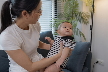 A young mother is holding her baby while spending quality time together indoors. She looks at her...