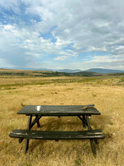 A weathered wooden picnic table with a lone white coffee mug sits in a golden grassy field, framed by rolling hills and a dramatic sky of clouds and blue