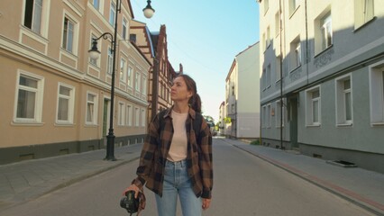 Light-skinned woman, 35 years old, holds camera while contemplating in serene urban street. Daytime...