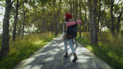 Fototapeta premium Young woman appears content while walking along paved path in serene park. Carrying yoga mat and backpack, she enjoys nature.