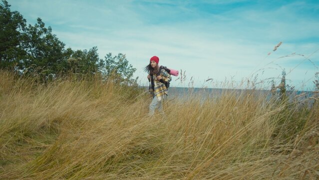 Young woman with backpack and walking sticks explores serene coastal grassland. Daytime adventure showcases peaceful nature. - Powered by Adobe
