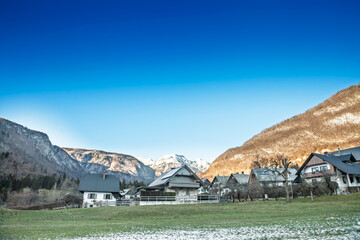 Snow-flecked chalets in Stara Fuzina sit beneath the Julian Alps, green meadow foreground and...