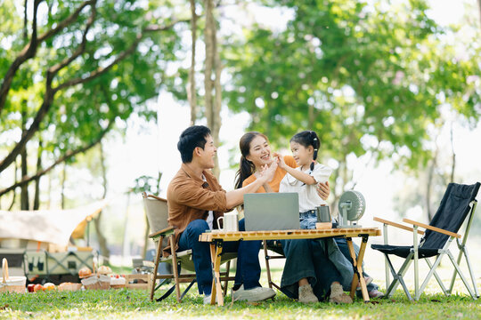 Joyful Asian family camping in a sunny garden, bonding over BBQ, tech, and laughter. Parents and daughter enjoy outdoor