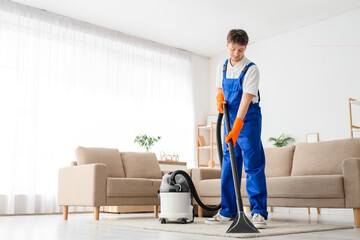 Young male janitor with modern vacuum cleaner cleaning carpet in living room