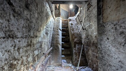 Underground passage stairway descent, cold dark basement. Eerie ancient staircase steps, steep handrail. Stone cut crumbling tunnel walls. Spooky cellar light, archaeological tomb ruins catacombs dig