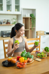 Asian woman holding green juice and apple, surrounded by fresh vegetables and salad in modern kitchen. Promotes healthy eating, fitness, and wellness lifestyle.
