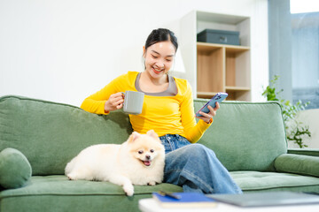 Asian woman using smartphone while relaxing on green sofa with fluffy dog and coffee mug. Cozy...