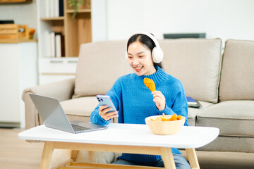 Asian woman using phone and laptop, wearing headphones, and eating fried chicken at home. Perfect for tech, lifestyle, snack, and remote work