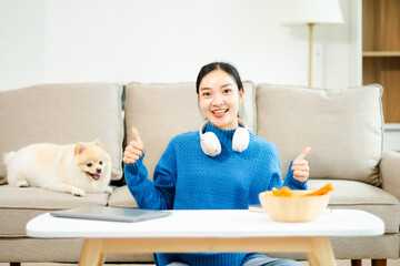 Young woman relaxing at home with wireless headphones, cute dog, snacks, and tablet. Cozy modern...