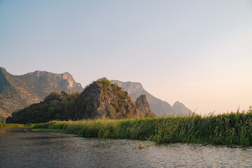 The mountain looms over a green landscape where a swamp meets a tranquil lake, water reflecting the...