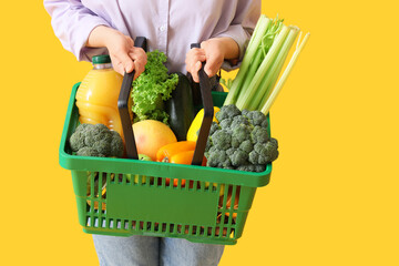 Fototapeta na wymiar Woman holding green shopping basket with different fresh products on yellow background